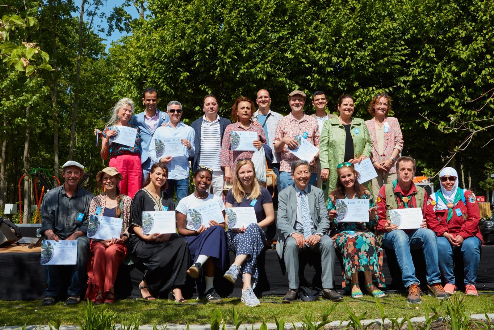 Groepsfoto van vrijwilligers van Huis van Vrede buiten in het groen, met certificaten van waardering